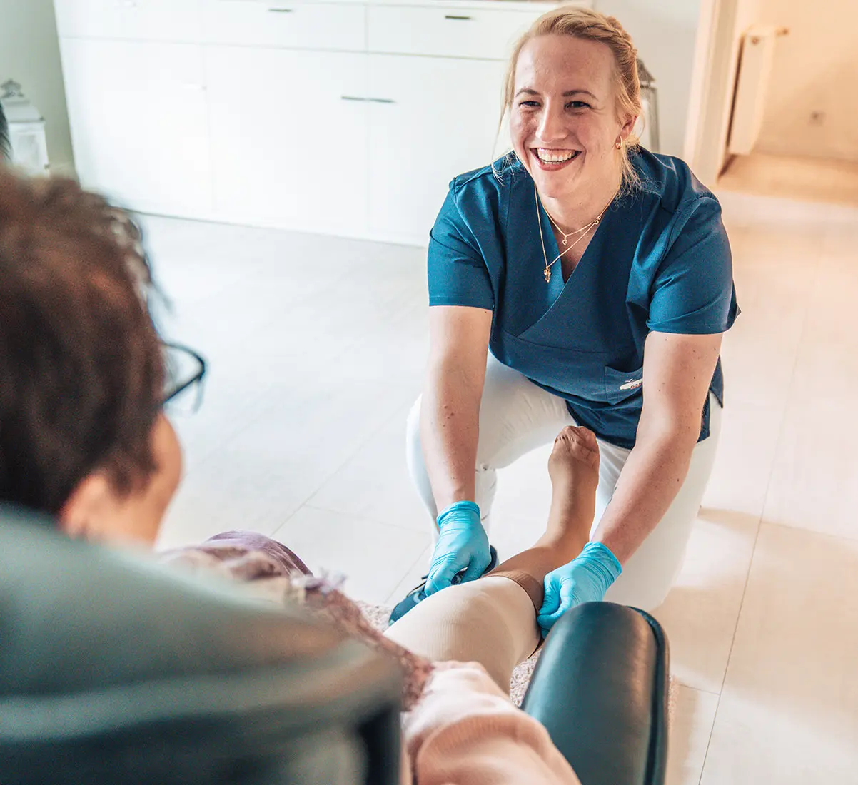 Pflegekraft in blauer Arbeitskleidung, die einer sitzenden Person mit einem Verband oder einer Bandage am Bein hilft, während sie freundlich lächelt.