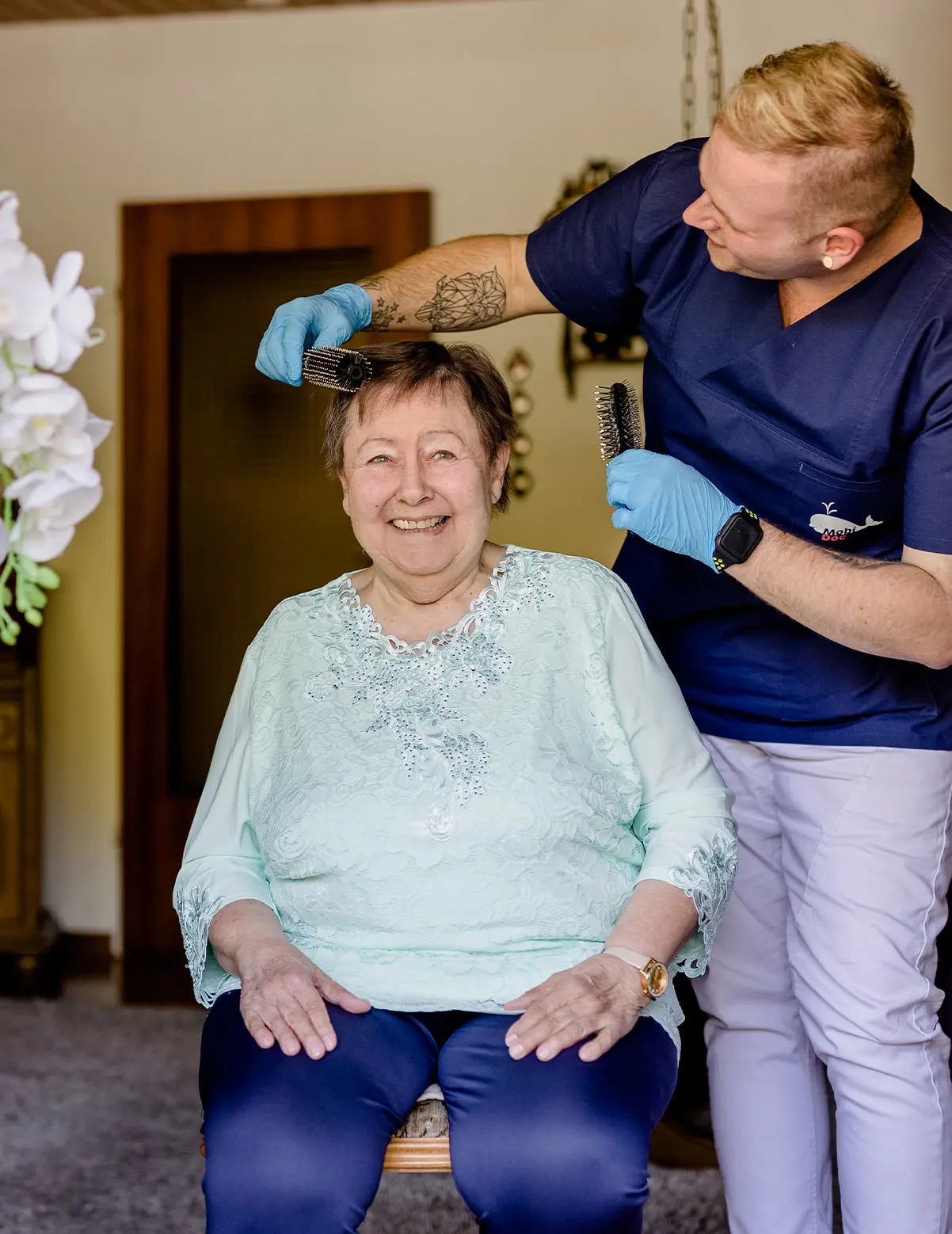 Pflegekraft in blauer Arbeitskleidung, die einer älteren Frau die Haare kämmt, während sie beide lächeln.