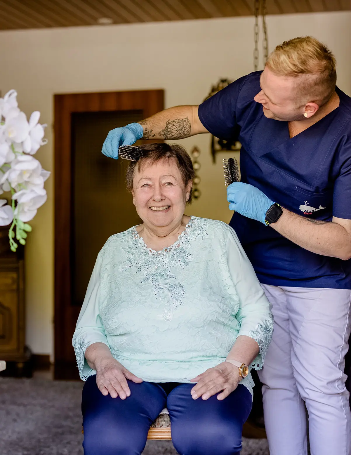 Pflegekraft in blauer Arbeitskleidung, die einer älteren Frau die Haare kämmt.